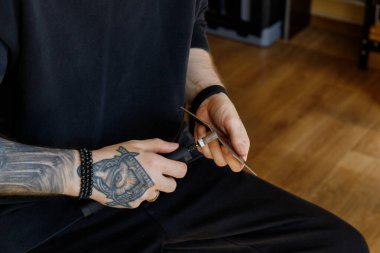 Leathercraft workspace with close-up of a tattooed hand using a rotary tool to polish the edge of a leather piece. Artisan seated in a dark outfit on wooden floor background. 