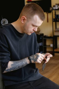 A close-up of a tattooed male artisan meticulously hand-stitching pieces of dark brown leather in a workshop. 
