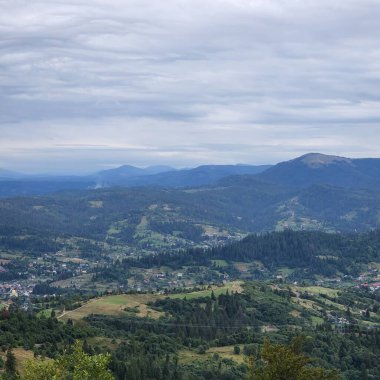 Dağlarla çevrili yeşil bir alanın panoramik görüntüsü ve bulutlu bir gökyüzü.
