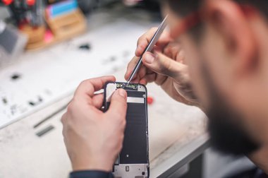 İş yeri üst görünümü, yakın plan. Bir elektronik tamir atölyesinde, tamirci akıllı telefonu tamir eder, cımbızı bir çok iş aletinden biri olarak kullanır.