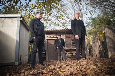 Üç şehirli ve yakışıklı sanatçılardan oluşan bir grup siyah şık takım elbiseli, açık havada poz veriyor.