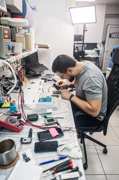 Electronics repair shop, a repairman is surrounded by tools and equipment. A technician repairs, cleans, controls a smartphone. Workplace top view, close-up.