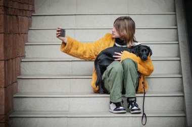 Sarı kürklü, sosyal medya etkileyicisi, siyah köpeğiyle şehrin merdivenlerinde oturan, akıllı telefon kullanan genç ve güzel bir kadın..