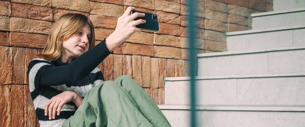 身穿黑白相间条纹毛衣的年轻漂亮女人 社交媒体的影响者 坐在建筑城的楼梯上 用智能手机 — 图库照片