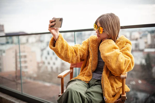 身穿黄色毛皮外套 戴着红色太阳镜的年轻漂亮姑娘坐在一座现代化建筑的阳台上 坐在一位导演的椅子上 使用智能手机 社交媒体影响者 摆姿势 背景城市 — 图库照片