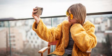 Sarı kürklü ve kırmızı gözlüklü güzel genç kız modern bir binanın terasında yönetici koltuğunda oturuyor. Akıllı telefon, sosyal medya etkileyicisi, fotoğraf çekme, selfie, poz verme, arka planda şehir kullanımı.