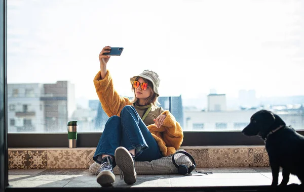 复制空间 漂亮的少女 互联网影响者坐在阳台的露台地板上 用智能手机 自私自利和娱乐 黄色皮大衣和红色眼镜 背景城市 — 图库照片