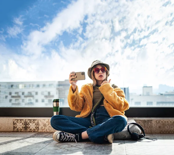 复制空间 漂亮的少女 互联网影响者坐在阳台的露台地板上 用智能手机 自私自利和娱乐 黄色皮大衣和红色眼镜 背景城市 — 图库照片