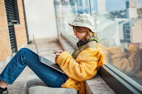 希斯特十几岁的女孩坐在阳台上 带着笔记本电脑 俯瞰城市 戴着帽子的年轻快乐女人在户外使用笔记本电脑 购物或上网工作 自由和自由职业者 — 图库照片