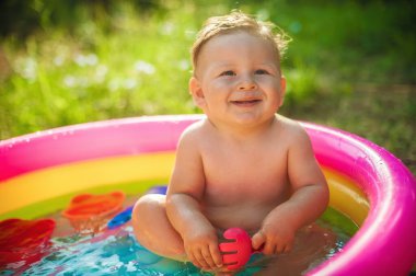 Küçük sevimli bir erkek bebek dışarıdaki çayır bahçesinde renkli bir havuzda banyo yaparken oyuncaklarla oynuyor. Yaz zamanı. Neşe ve mutluluk. Aşk ve aile 