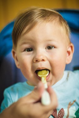 Beautiful baby boy eating porridge food, with a spoon. Hungry baby with open mouth enjoying food. Mother feeding her baby