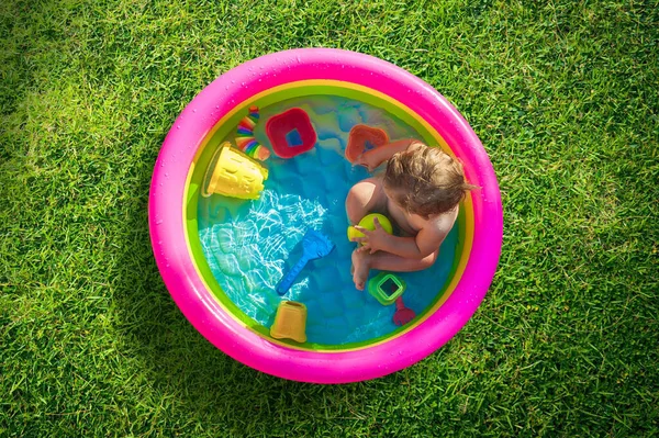 Baby boy sitting in a pool at the garden and playing with water toys. Hot summer day in nature
