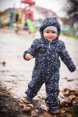 Küçük çocuk yağmurun ortasında duruyor ve yağmurlu bir günde şehir sokaklarında yağmur birikintileriyle oynuyor. Yağmurla ıslanmış macera