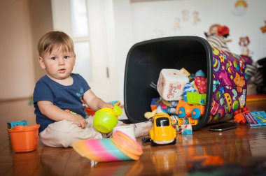 Genç bir çocuk hareketli bir oyun odasında yerde oturuyor, devrilmiş bir çöp kutusundan dökülen bir oyuncak hazinesini keşfediyor, etrafa neşe saçıyor..