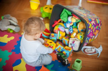 Genç bir çocuk, güzel bir dizi oyuncakla çevrili, parlak, desenli bir paspasın üzerinde oturur. Oda doğal ışıkla dolu. Küçük olan eğlenirken, neşeli bir atmosfer yaratıyor..