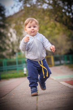 Neşeli, gri ve mavi giyimli bir erkek bebek, enerjik bir şekilde parkın asfalt yolu boyunca koşuyor, küçük ayakları etrafındaki dünyayı keşfetmeye hevesli..