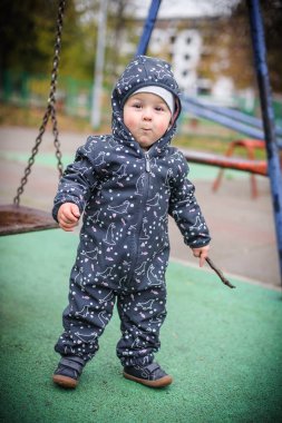 Sıcak bir şekilde giyinen erkek bebek, elinde sopayla salıncakta sallanırken oyun sahasında serin sonbahar havasının tadını çıkarıyor. Merakı parlıyor.