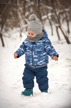 Rahat bir kışlık ceket ve şapka giymiş neşeli genç bir çocuk, kar kaplı manzarada zevkler, pofuduk beyaz battaniyenin içinde küçük adımlar atarken gülümsüyor..