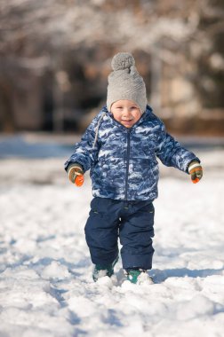 Mutlu genç bir çocuk kış kıyafetleri içinde, büyük bir şapka ve renkli eldivenlerle karlı bir tarlada dolaşır. Güneş ışığı huzurlu kış manzarasını aydınlatır..