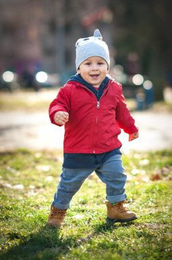 Parlak kırmızı ceketli neşeli bir çocuk güneşli bir parkta çimenli bir alanda geziniyor. Bulaşıcı gülümsemesi çocukluk maceralarının neşesini yakalıyor..
