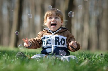 Gülümseyen bir çocuk, sakin bir ormandaki yemyeşil çimenlerde oturur. Etrafı güneş ışığında parıldayan kabarcıklarla çevrilidir. Saf neşe saçar..