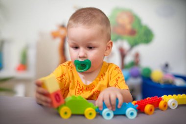 Yürümeye başlayan çocuk, oyuncaklarla dolu neşeli, aydınlık bir oyun odasında, masanın üzerine bir tren seti inşa ederek, canlı plastik yapı taşlarıyla meşgul oluyor..