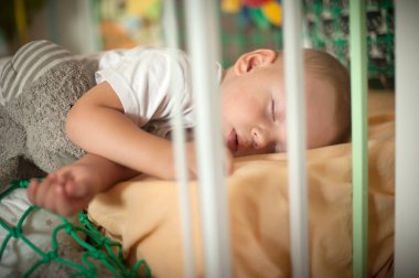 Baby lies comfortably asleep on a soft blanket in a crib, with gentle sunlight illuminating the vibrant nursery, creating a serene atmosphere perfect for rest. clipart