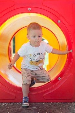 Küçük bir çocuk renkli bir tünelden çocuk parkına tırmanıyor. Sahne canlı, sarı ve kırmızı renkler, güneşli bir öğleden sonra dışarıda eğlenmek için mükemmel..