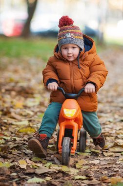 Sıcak turuncu bir ceket ve renkli bir şapka giyen küçük bir çocuk, yapraklarla kaplı bir patikada parlak turuncu bir bisiklete biniyor. Neşeli atmosfer güneşli bir sonbahar gününü yansıtır..