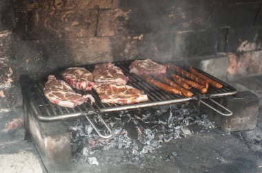 Steaks and sausages are being grilled over glowing coals in a traditional stone barbecue. Smoke swirls around as the meat cooks, creating a flavorful aroma on a warm summer evening. clipart