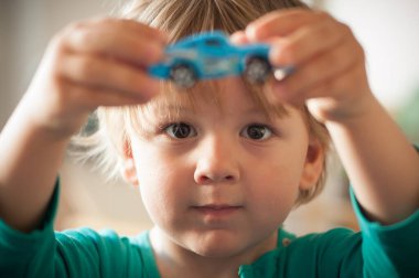 Açık kahverengi saçlı genç bir çocuk küçük mavi bir oyuncak arabayı tutuyor, dikkatle ona bakıyor. Yumuşak doğal ışık iç mekanı doldurur ve çocuğun parlak ifadesini güçlendirir..