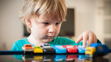 Genç bir çocuk, parlak bir iç mekan ortamında merak ve konsantrasyon gösteren yansıtıcı bir yüzeyde oyuncak araba koleksiyonu inceler ve organize eder..