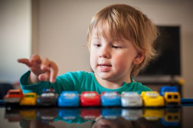 Genç bir çocuk, renkli oyuncak arabalardan oluşan bir dizilimle meşgul, öğleden sonraları kapalı alanda konsantrasyon ve neşe sergiliyor..