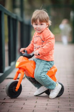 Küçük bir çocuk parkta bir patika boyunca turuncu bir oyuncak motosiklet sürüyor. Çocuk gündelik giysiler giyiyor ve açık hava deneyimini seviyor, mutlu bir şekilde gülümsüyor..