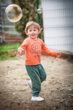 Turuncu ve yeşil kıyafetli gülümseyen bir çocuk, parlak, güneşli bir öğleden sonra kumlu bir parkta yüzen baloncuklardan sonra heyecan içinde koşar..