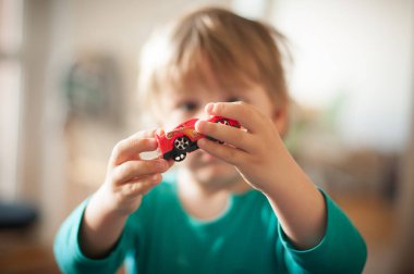 Açık kahverengi saçlı bir çocuk, doğal ışıkla dolu parlak bir odada otururken yüzünde kırmızı bir oyuncak araba tutuyor, gülümsüyor. Atmosfer neşeli ve sıcak..