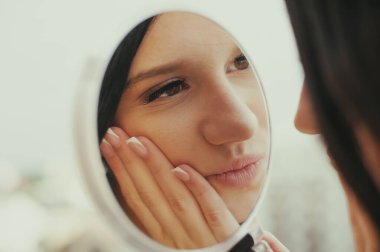 Siyah saçlı genç bir kadın el aynasına bakıyor, yüzüne dokunuyor. Muhtemelen görünüşünü kontrol ediyor ya da sosyal medya için poz veriyor, şık bir görünüm sergiliyor..