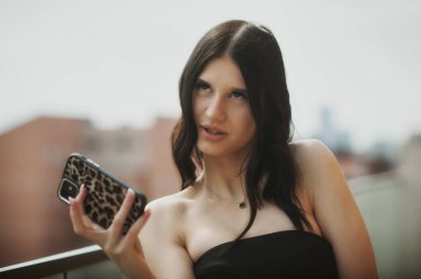 Şık bir kadın balkonda otururken telefonu tutar. Düşünceli bir şekilde uzak diyarlara bakıyor, berrak gökyüzünün altında canlı bir şehirle çevrili..