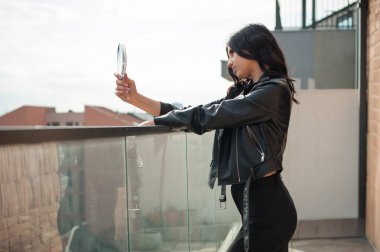 Gündüz vakti genç bir kadın balkonda dikiliyor. O, yüzüne bakmak için küçük bir ayna tutarken siyah bir deri ceket ve etek giyiyor..