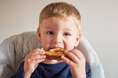 Sarışın, açık tenli bir çocuk ızgara peynirli sandviç yiyor. O, sandalyede otururken iki eliyle sandviçi tutuyor, yüzünde neşeli bir bakışla çiğniyor..