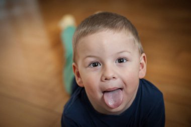 Kısa sarı saçlı küçük bir çocuk ahşap bir zeminde yatıyor. Çocuk koyu mavi bir gömlek giyiyor. Kameraya bakıyor ve şakayla dilini dışarı çıkarıyor..