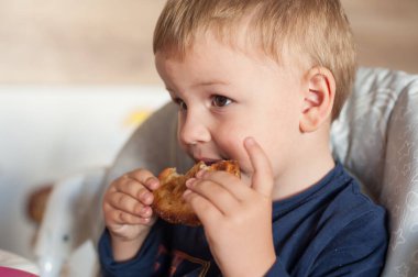 Sarı saçlı, açık tenli bir çocuk bir sandalyede oturmaktadır. Aktif olarak bir parça yemek yiyor, iki eliyle tutarak bir ısırık alıyor..