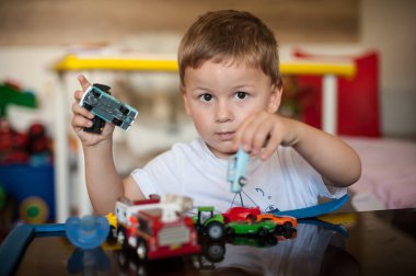 Kahverengi saçlı genç bir çocuk oyuncak arabalarla, bir traktörle ve kapalı bir masada itfaiye arabasıyla oynuyor. Elinde oyuncak araçlar tutuyor ve ileriye bakıyor..