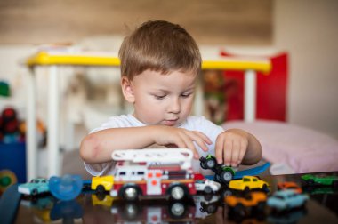 Beyaz tişört giyen sevimli genç bir çocuk oyuncak arabalarla ve yansıtıcı bir masada oyuncak bir itfaiye arabasıyla oynuyor. Oyun odası parlak ve renklidir. Arka planda diğer oyuncaklar görünür..