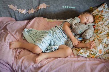 Genç bir çocuk, yumuşak bir yatakta, renkli yataklarla çevrili bir şekilde mışıl mışıl uyuyor. Sıcak atmosferde doldurulmuş bir hayvan var. Huzurlu bir an yaratıyor..