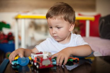 Canlı oyuncak arabalar ve birkaç oyuncakla çevrili bir masada oturan küçük bir çocuk. Çocuk meşgul, dikkatlice oynuyor ve oyuncakları düzenliyor. Eğlenceli ve hayat dolu bir atmosfer odayı dolduruyor..