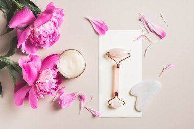 Patenli ve gua sha gül kuvars, krema ve pembe şakayık çiçekleri. Spa konsepti, yüz bakımı.