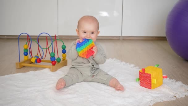 赤ちゃんは床に座り ポップに噛み付いたり シンプルに薄暗くなったりします 周りのさまざまなおもちゃ — ストック動画