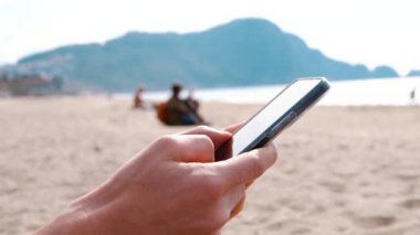 Bir adamın eli telefonu tutar. Arka planda deniz ve kumsal.