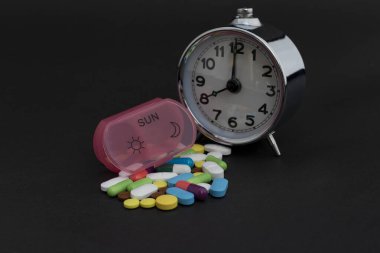 Alarm clock with a pill organizer box and pills on black background. clipart